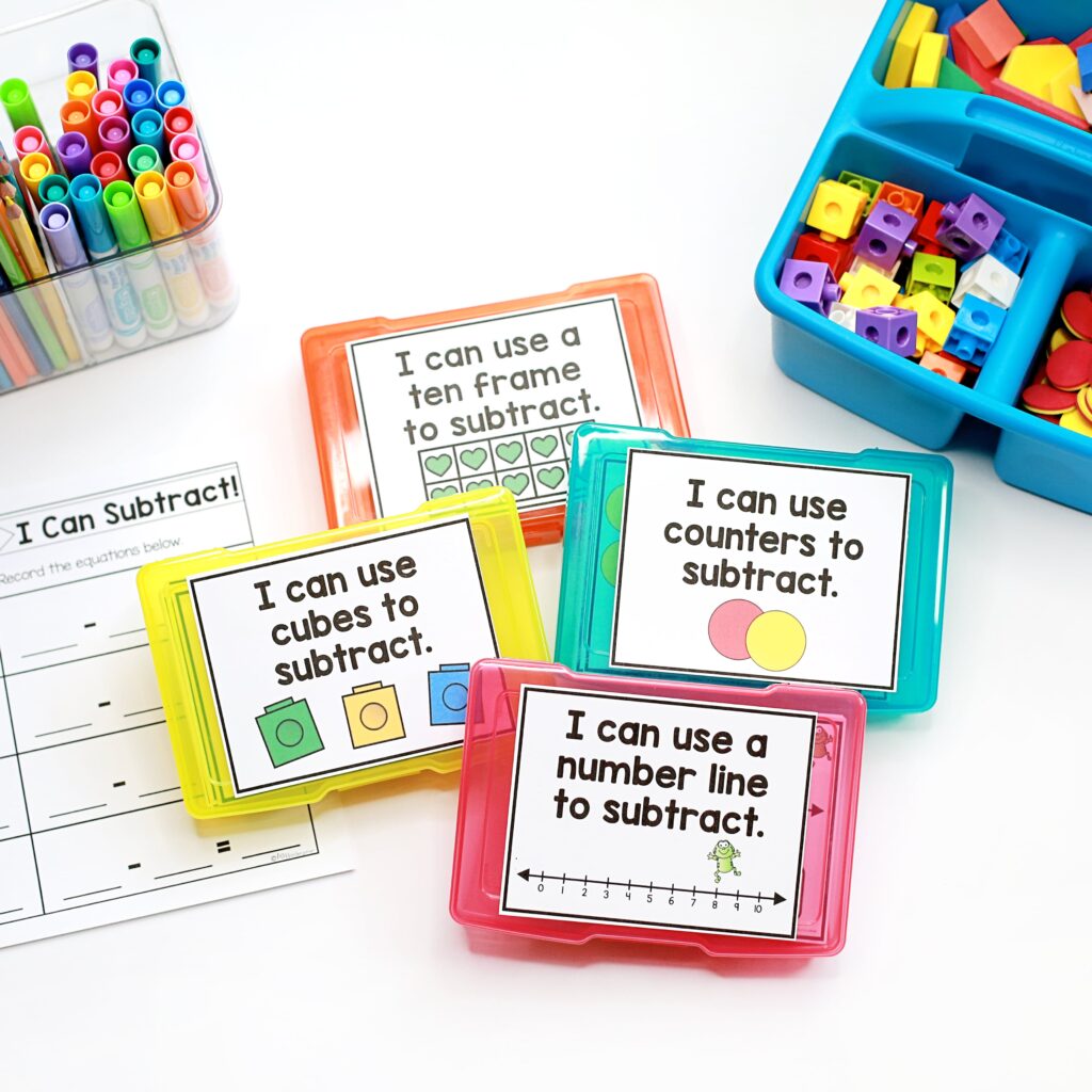 A white desk top contains four sets of subtraction task cards, a bin of math manipulatives, a container of markers, and a subtraction recording sheet.