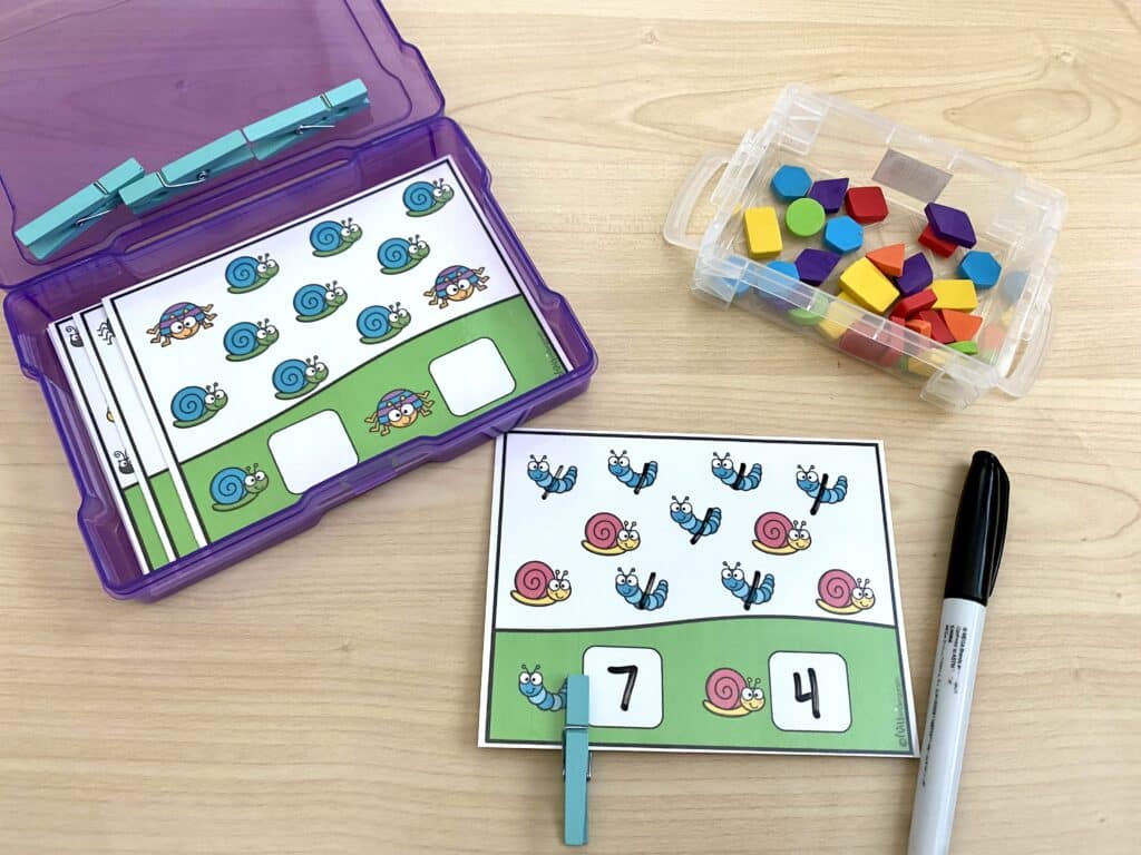 A counting task card is being completed, with a clothes pin and dry erase marker. A bin of small shape manipulatives is also on the desk.