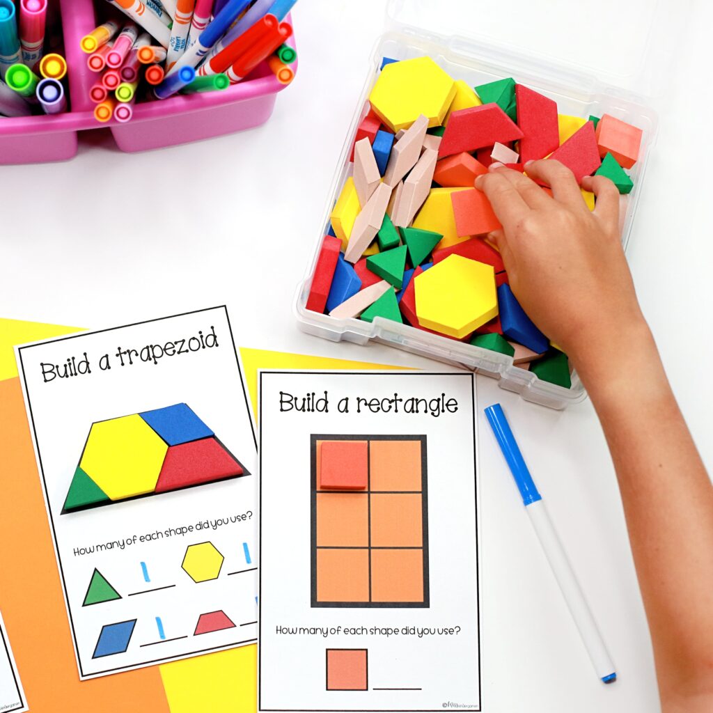 Two shape building cards are in use, with a hand choosing pattern blocks from a container to build the larger shapes.