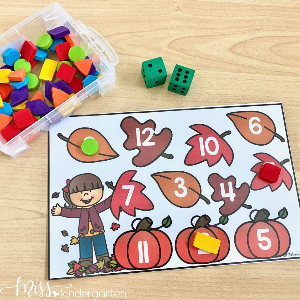 A fall-themed roll and cover game is being used with shape manipulatives and two green dice.